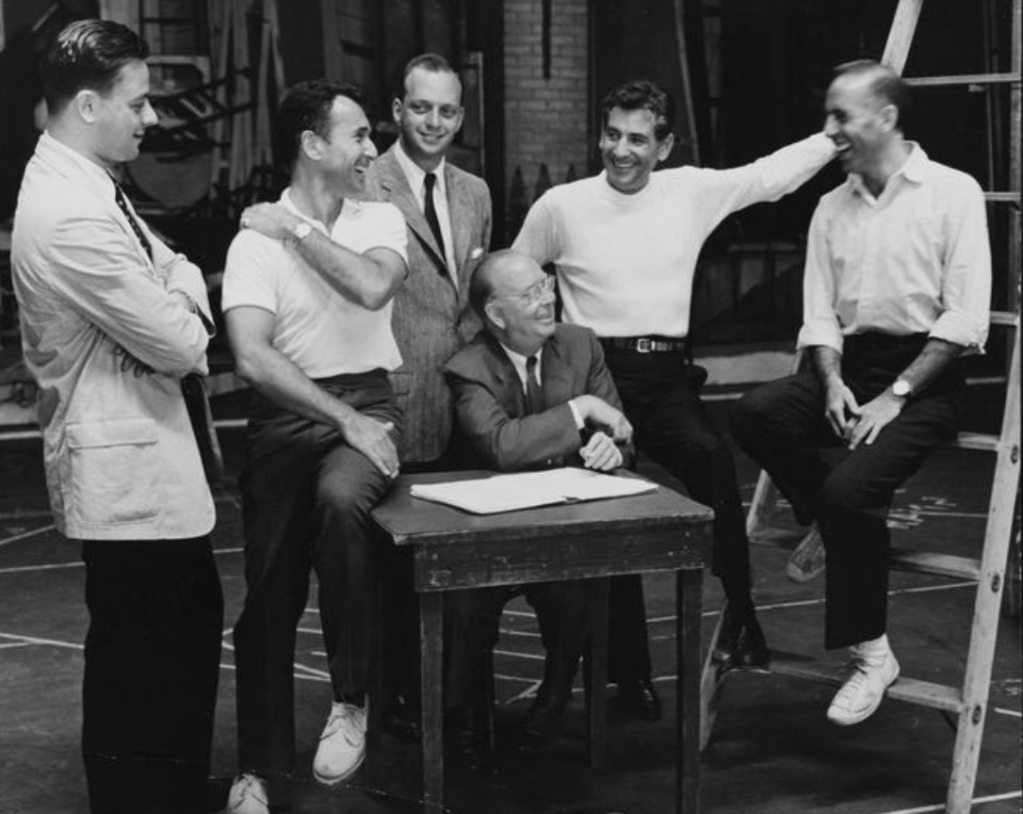 Robert E. Griffith sites at a desk surrounded by the creators of "West Side Story," who are enjoying a laugh.